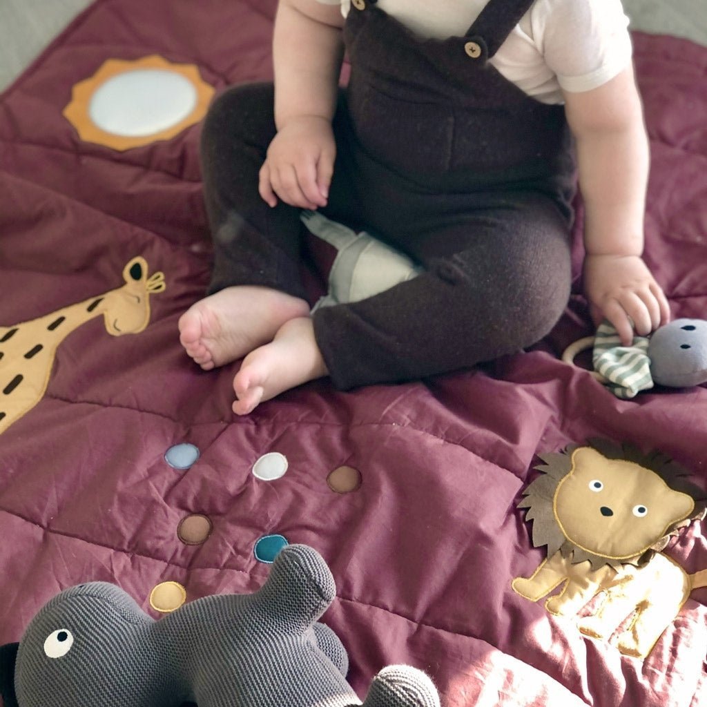 Een baby zit met gekruiste benen op de Nuuroo Alin activiteitenmat, een bordeauxrode speelmat met dierenillustraties, draagt een bruine overall en houdt een knuffel vast terwijl hij geniet van het zonlicht en zijn motorische vaardigheden ontwikkelt.