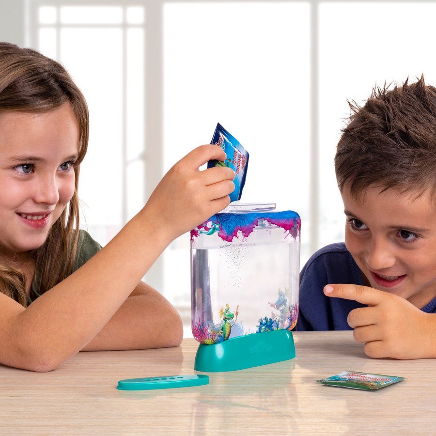 Twee lachende kinderen zitten aan een tafel en kijken opgewonden terwijl de een een zakje toevoegt aan een klein, kleurrijk aquarium gevuld met water en plastic zeedieren. Een helder raam bevindt zich op de achtergrond.