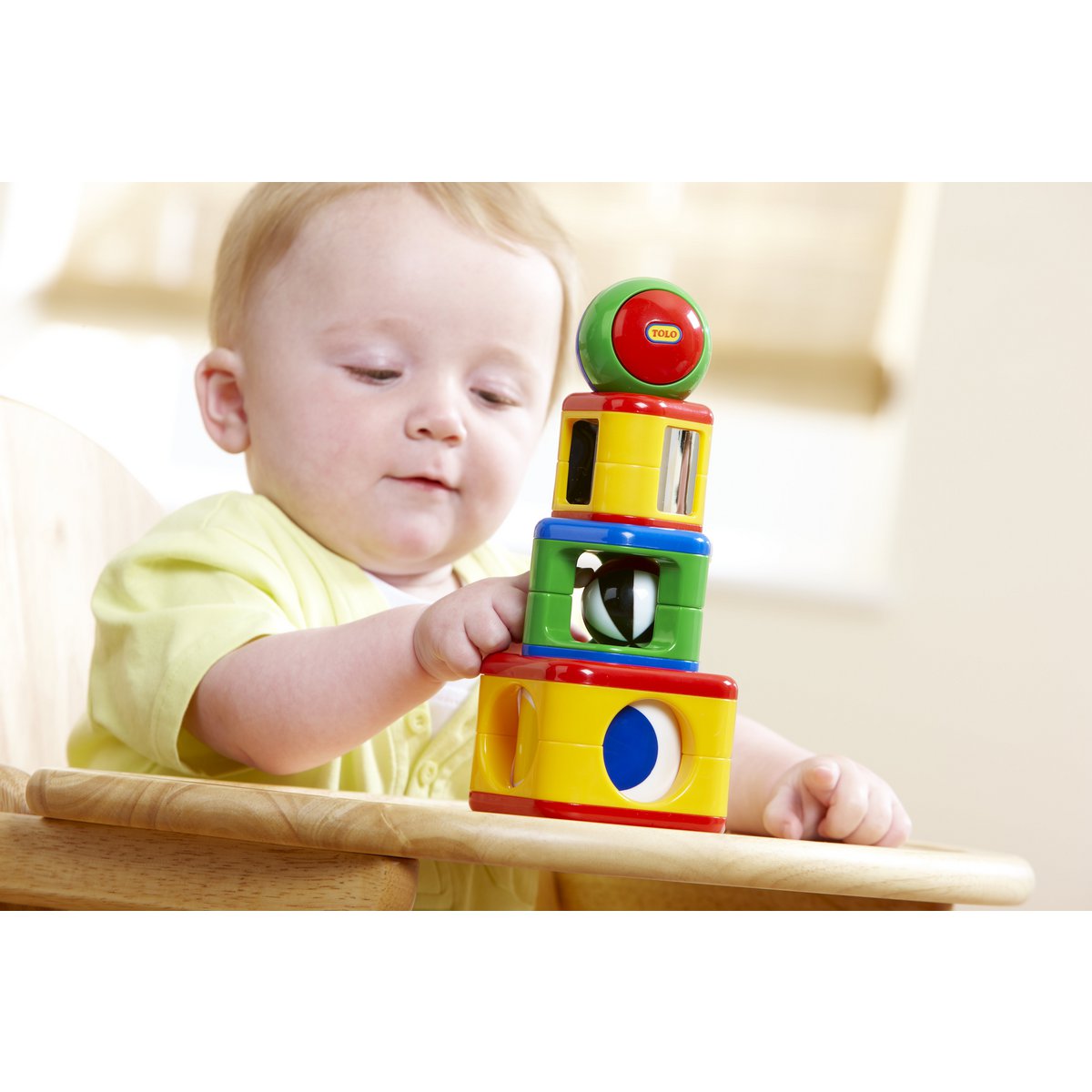 Een lichtharige baby in een geel shirt zit in een kinderstoel en stapelt de Tolo Classic Activiteitenspeelgoed Stapeltoren - 4-delig van Tolo, met kleurrijke stukjes structuur en een groene bal bovenop. De zachte achtergrond benadrukt het ontwikkelen van vaardigheden.