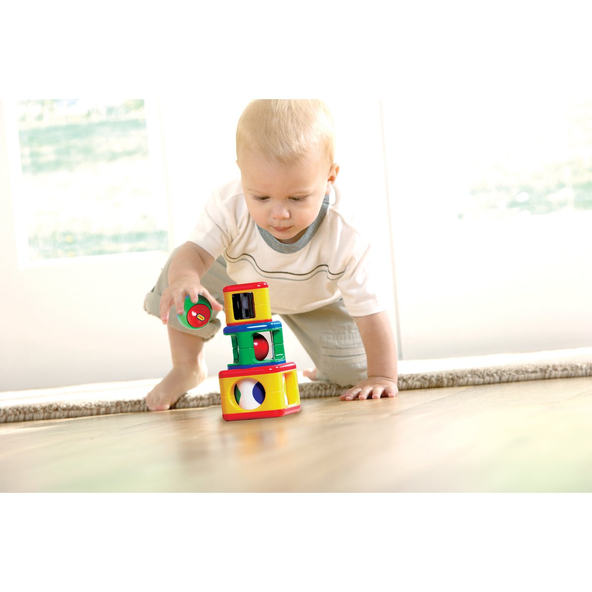Een baby met kort blond haar, in een wit shirt en grijze korte broek, knielt op een houten vloer en speelt met de Tolo Classic Activiteitenspeelgoed Stapeltoren - 4-delig. Het heldere Tolo speelgoed heeft patronen en ronde ramen waarachter zonlicht naar binnen stroomt.