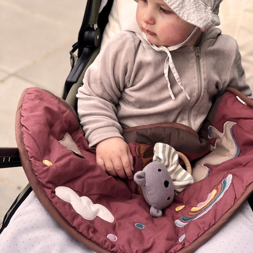 Een baby met een lichtgrijze capuchon en zonnehoed zit in een kinderwagen op de Nuuroo Vilma speelmat, gewikkeld in een kastanjebruine deken met wolken en regenbogen, met een zacht grijs speeltje in de hand en een bedachtzame blik.
